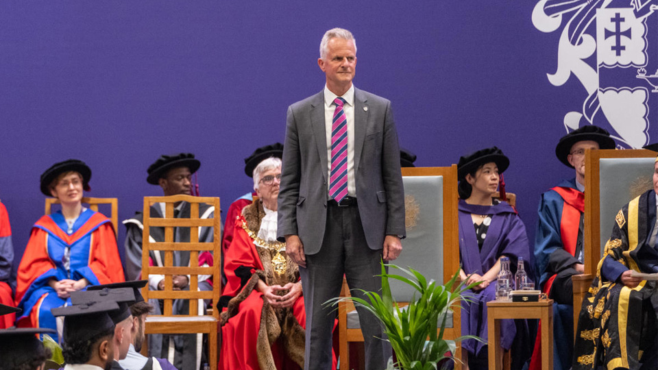 A man in a suit stands on stage during a formal ceremony; others in academic regalia are seated behind him against a purple backdrop.